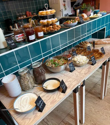 Uitgebreid brunchbuffet met taartenstandaard, donuts, confituur en broodmandjes tegen de blauwgroen betegelde wand van Pepper